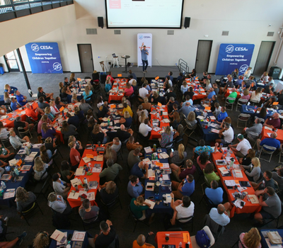 Past Summer Leadership Retreat Photo showing many tables with different colored table clothes, and around 75 people all in frame sitting at the tables from above. Ted is in the front on a stage, and everyone is very engaged and looking at Ted on stage. 