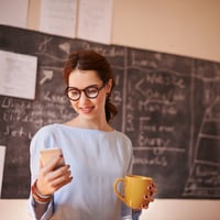 Teacher checking her phone in a classroom. Background includes a chalkboard covered in math equations, showing she is actively in her classroom teaching